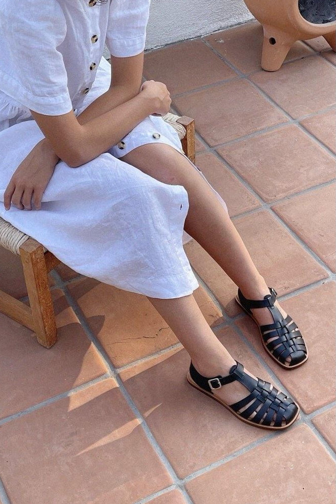 LOQ Rio sandal in black worn with white dress seated on tile floor showing fisherman sandal styling and buckle strap