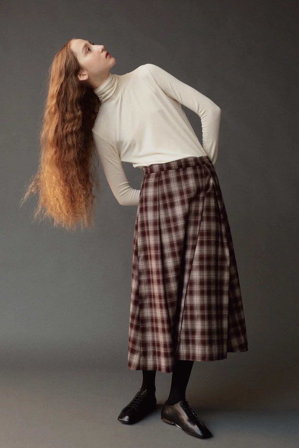 Model leaning to the side in the Mijeong Park brown plaid midi skirt and ivory turtleneck
